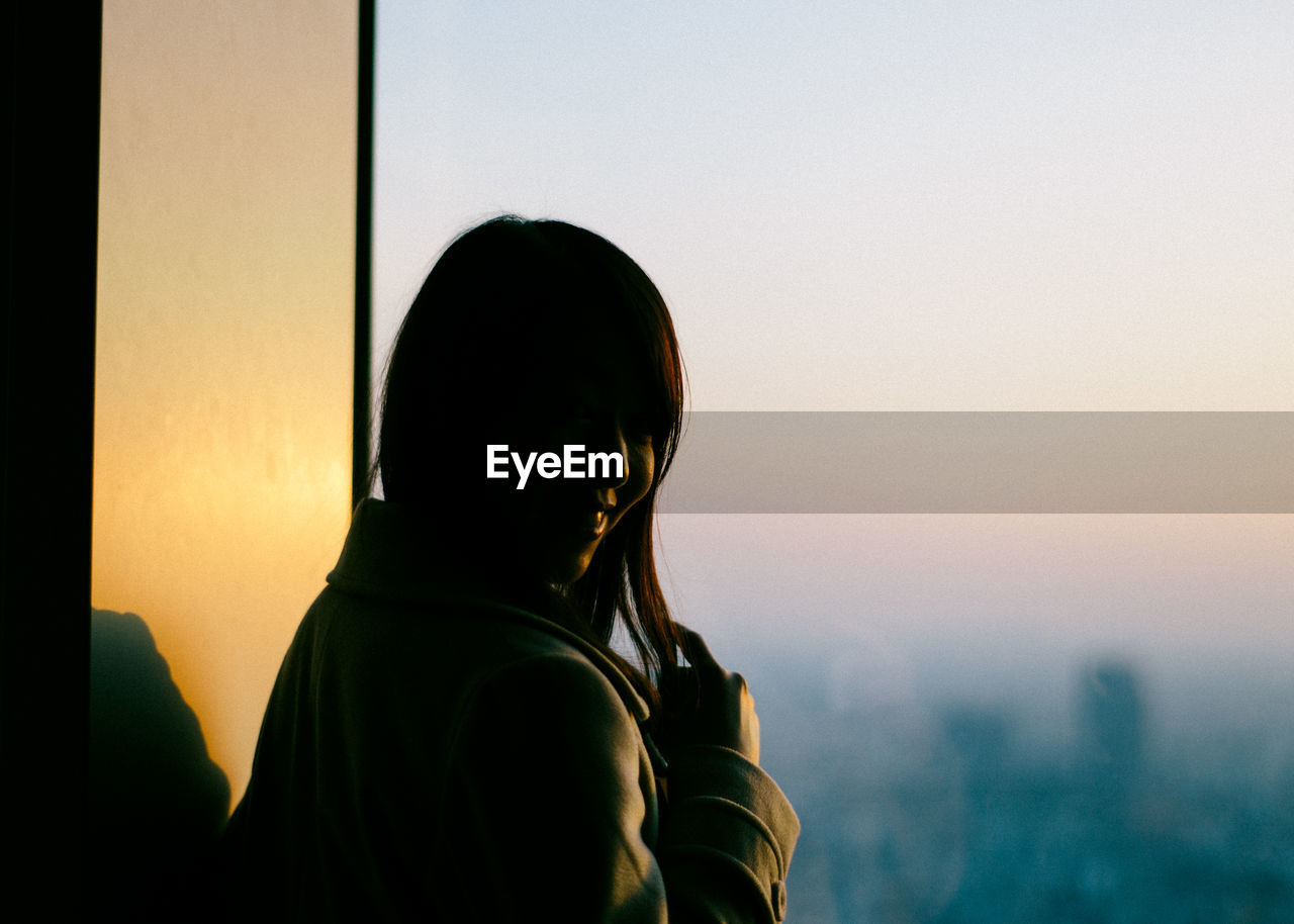 Woman standing by window during sunset