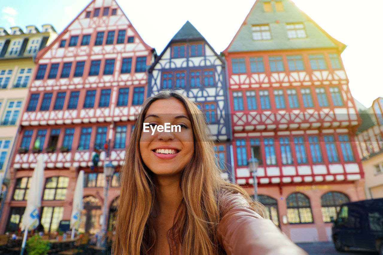 PORTRAIT OF SMILING YOUNG WOMAN AGAINST BUILDINGS