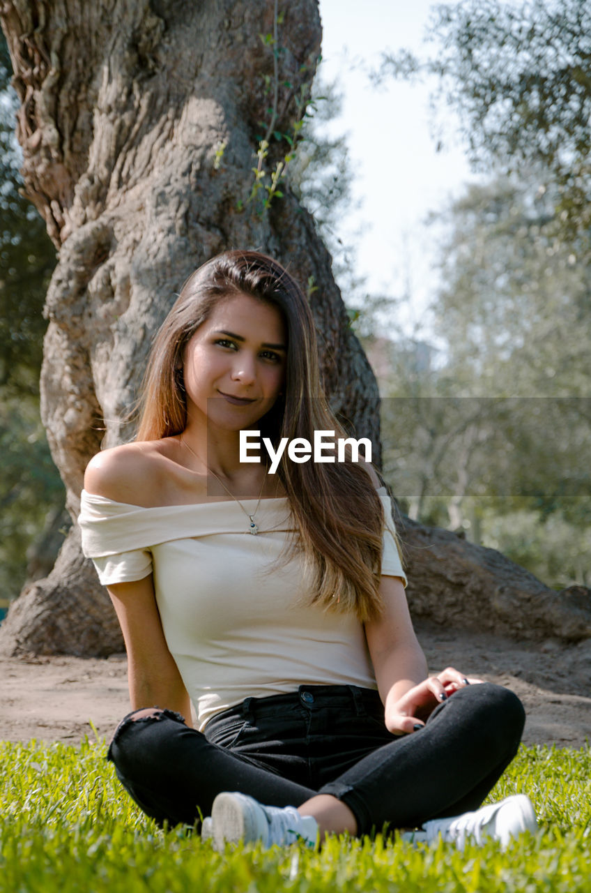 Full length portrait of smiling young woman sitting against trees in park