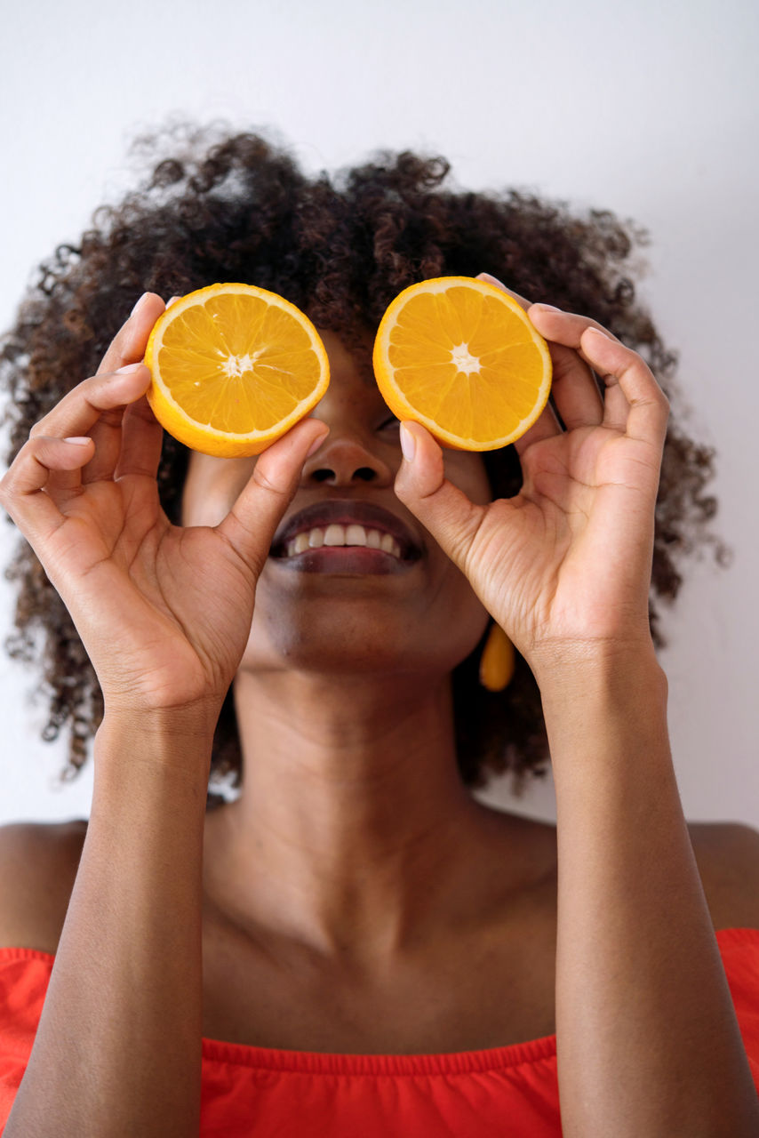 Black woman with afro hair holding cut orange halves in front of her eyes having fun