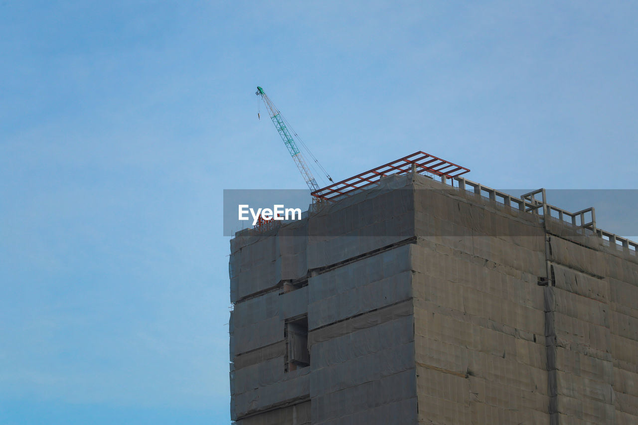 LOW ANGLE VIEW OF CRANE BY BUILDING AGAINST BLUE SKY