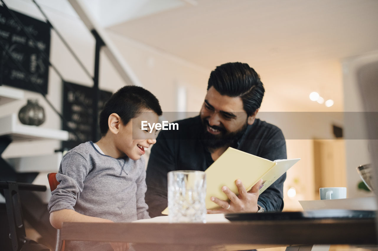 Smiling father teaching autistic son while sitting at table in house