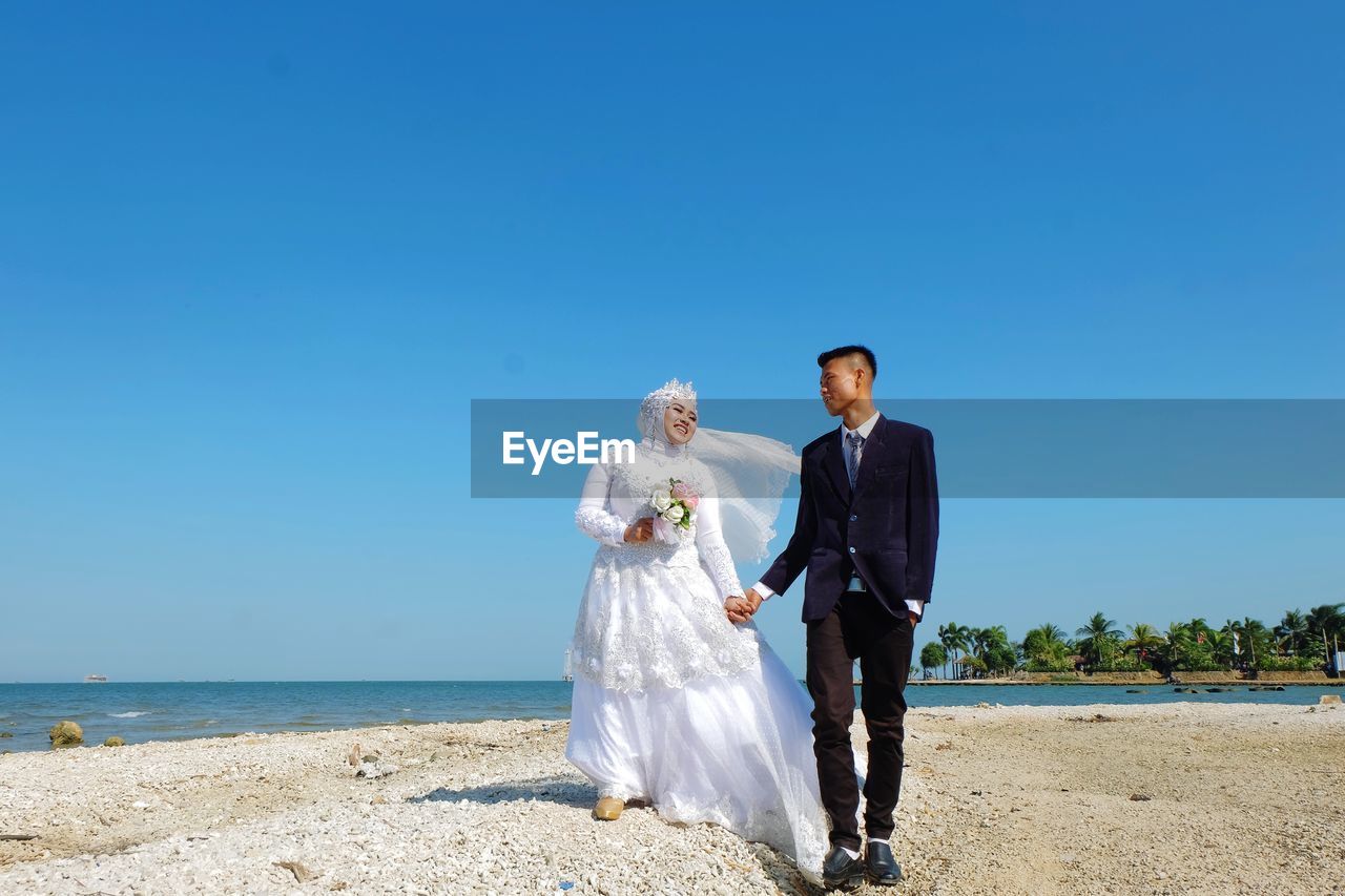 Couple holding umbrella while standing on shore against blue sky