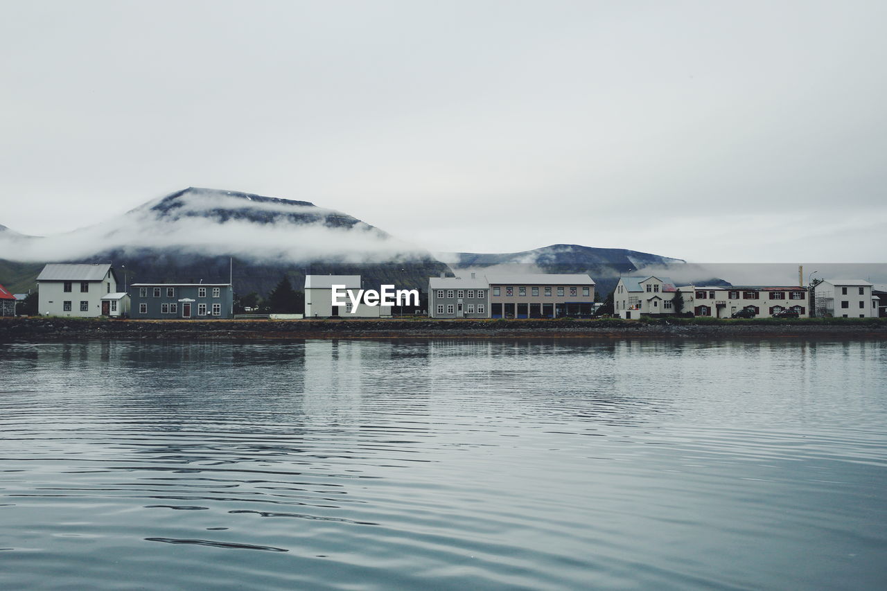 Houses on riverbank by mountains against sky