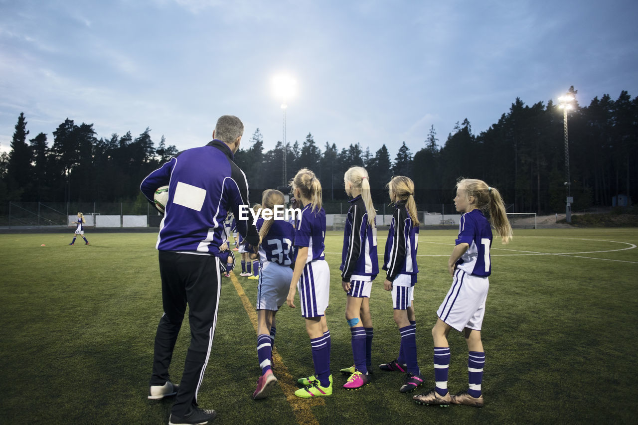 Coach explaining female soccer team on field against sky