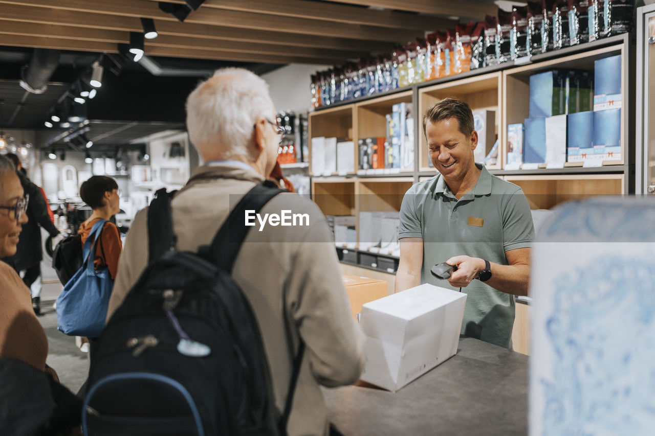 Smiling sales clerk scanning appliance box while customers at checkout counter in electronics store