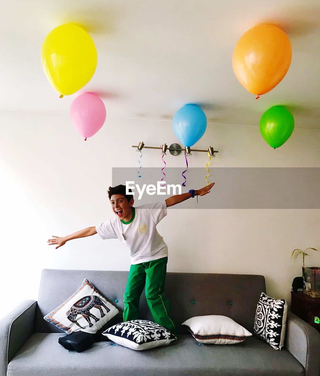 Portrait of boy standing by colorful balloons on sofa in living room at home