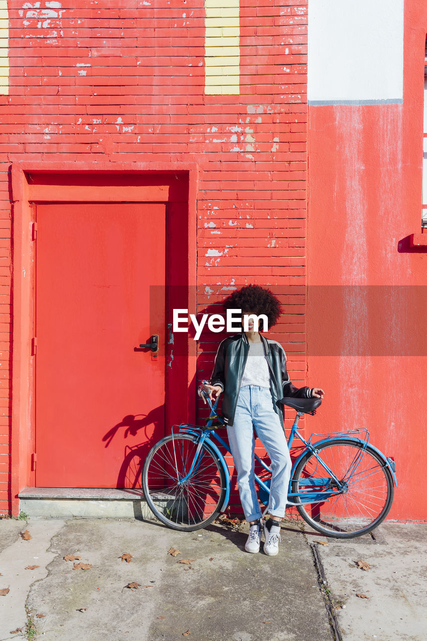 Smiling young woman leaning on bicycle in front of red building