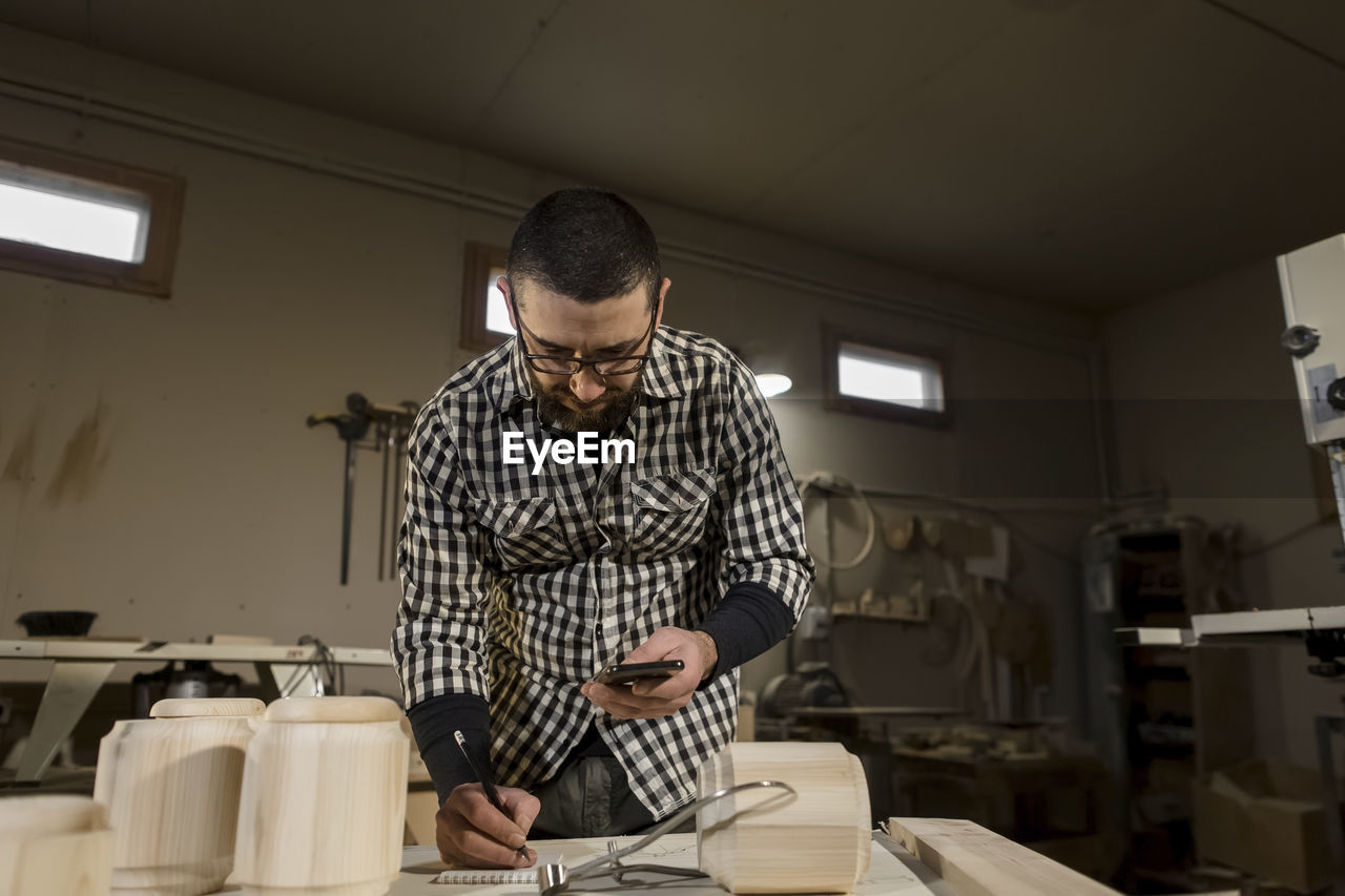 Carpenter holding smart phone writing in note pad at workshop