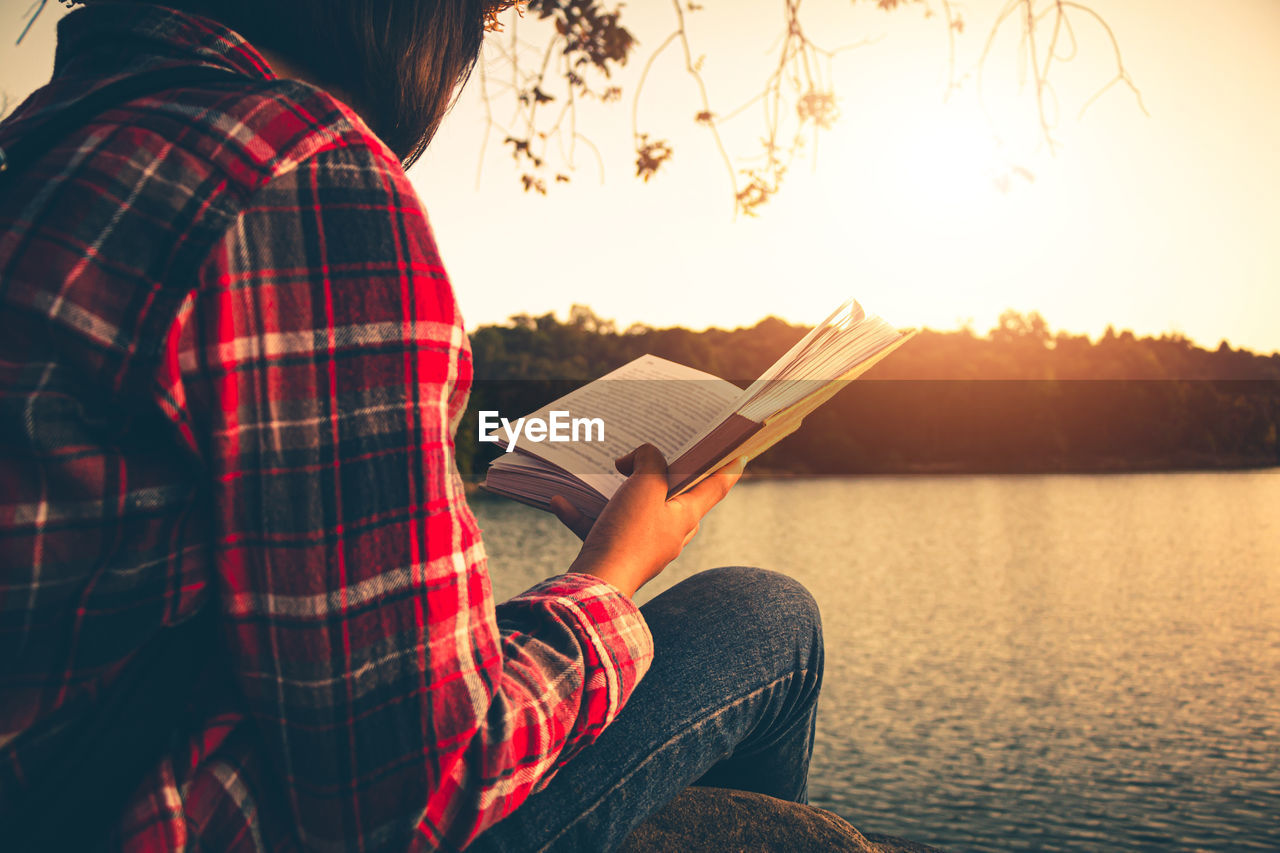 Man holding book while sitting by lake against sky during sunset