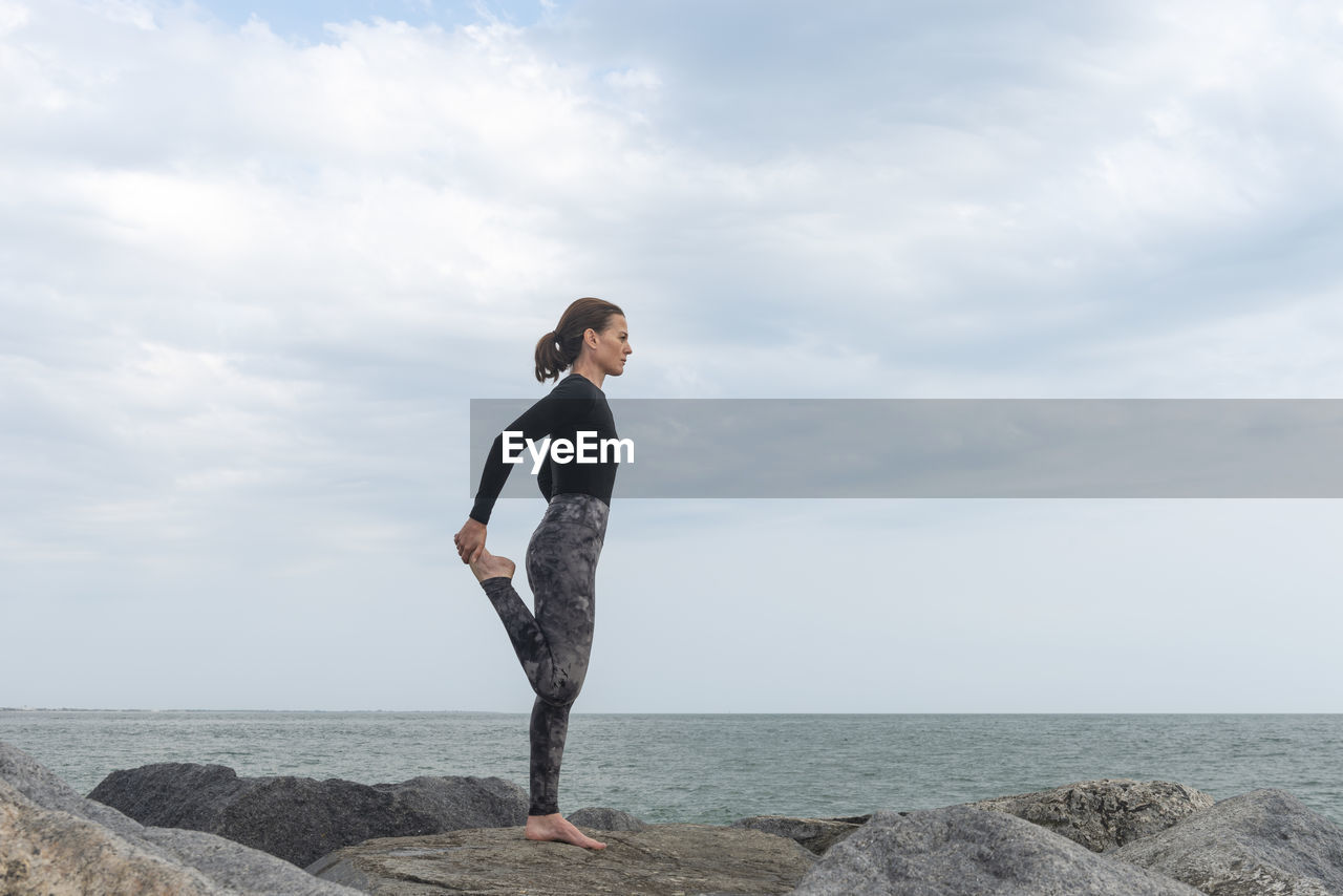 Sporty female doing a leg stretch, warm up exercises by the sea.