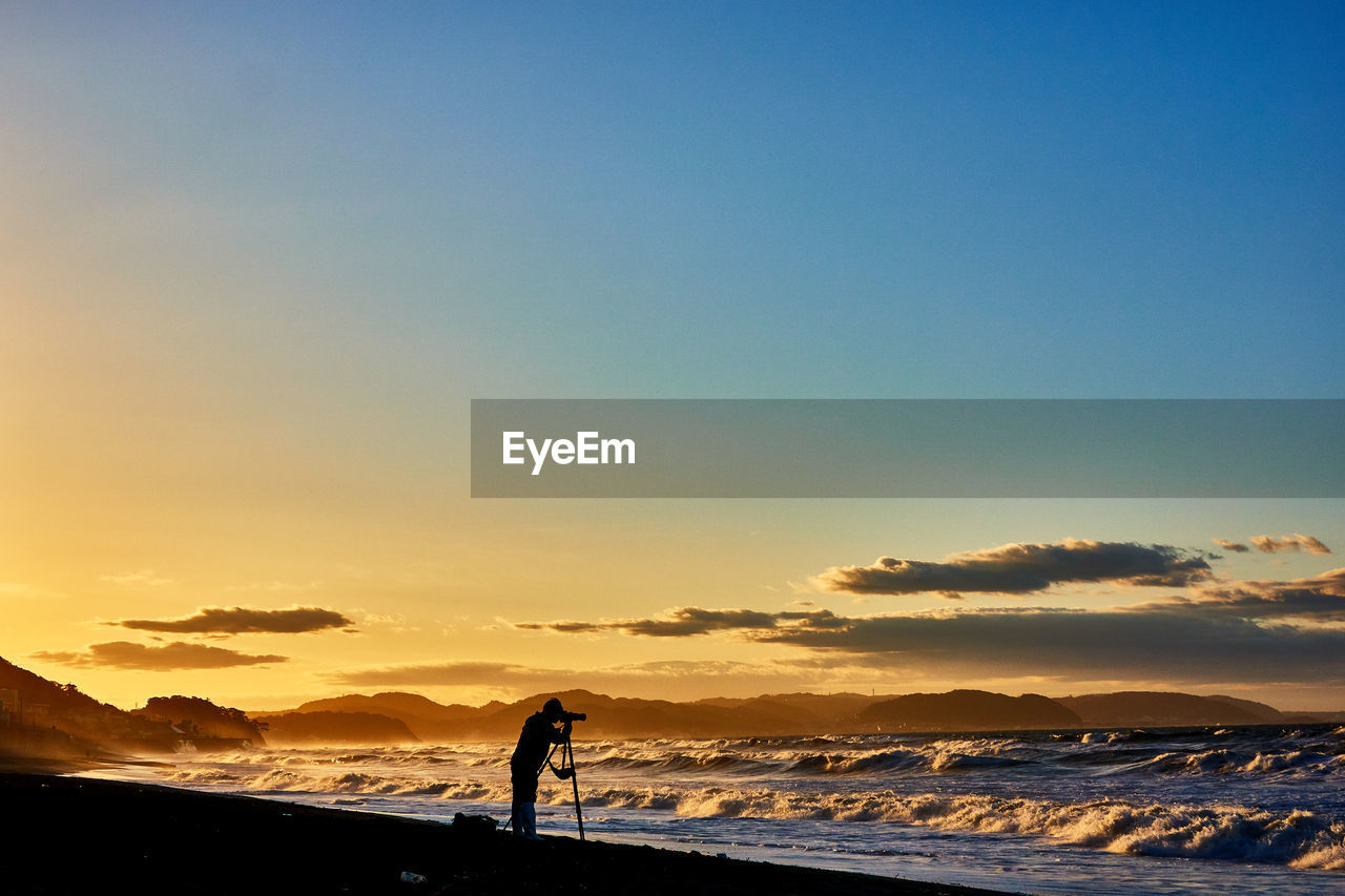 Scenic view of sea and standing person against dramatic sky during sunrise