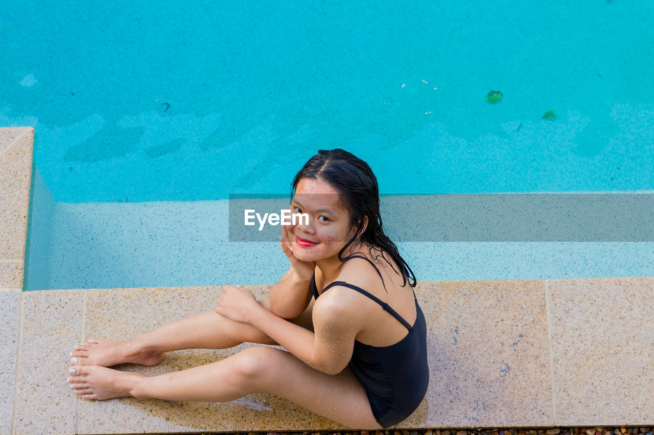 Beautiful asian woman sitting at pool side on sunny day while smiling