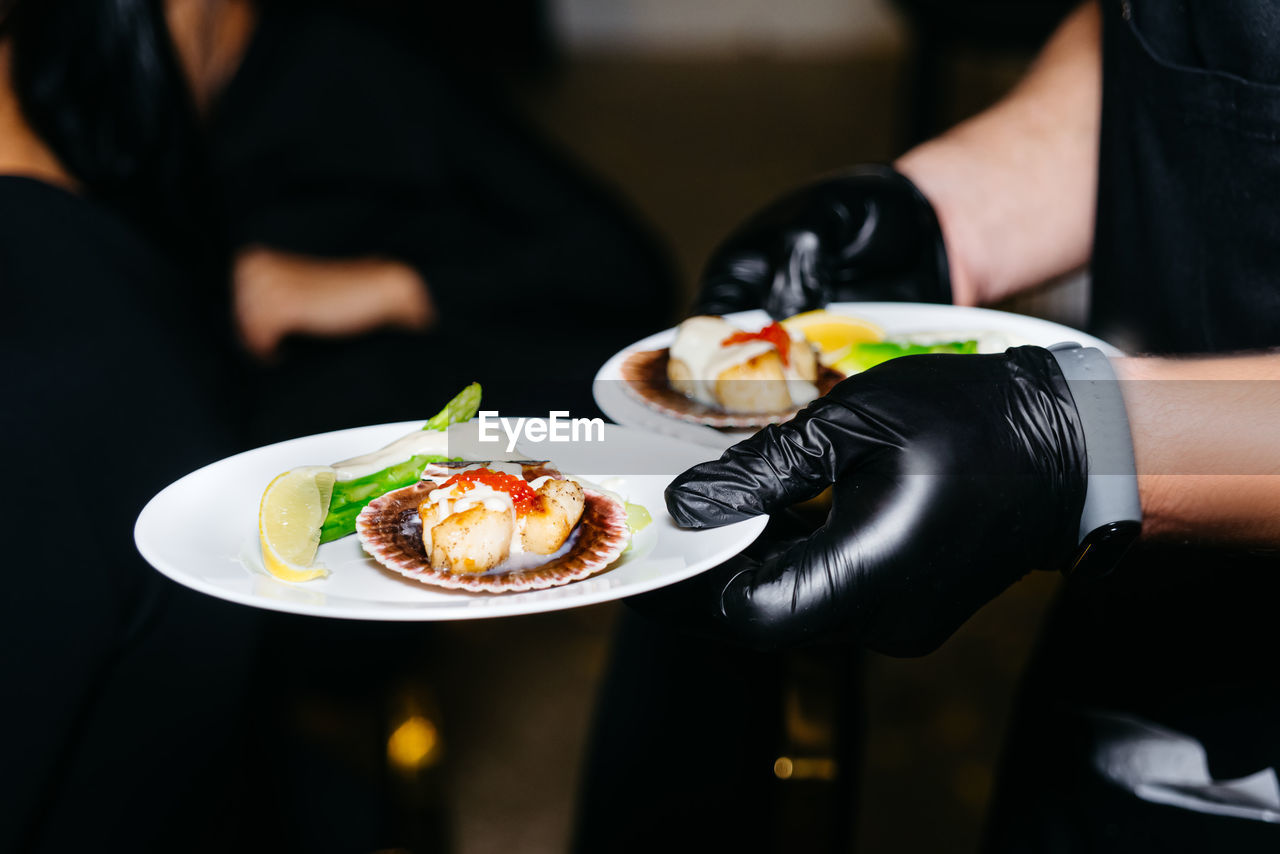 Close-up of waiter's hands in black gloves serving scallops. two portions of haute cuisine, catering