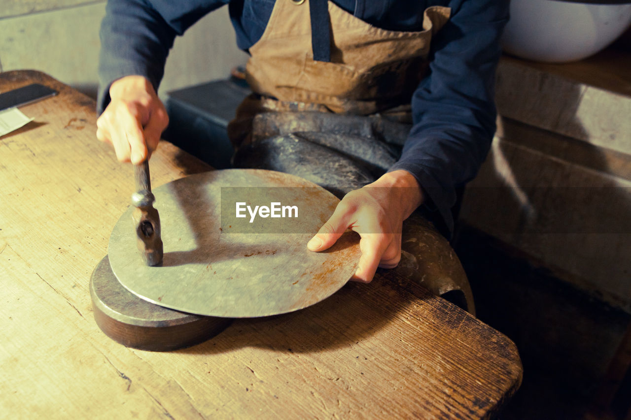 Midsection of worker hammering metallic plate in workshop