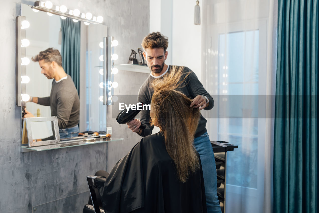 Ethnic male hairdresser drying hair of female client with closed eyes in modern beauty studio
