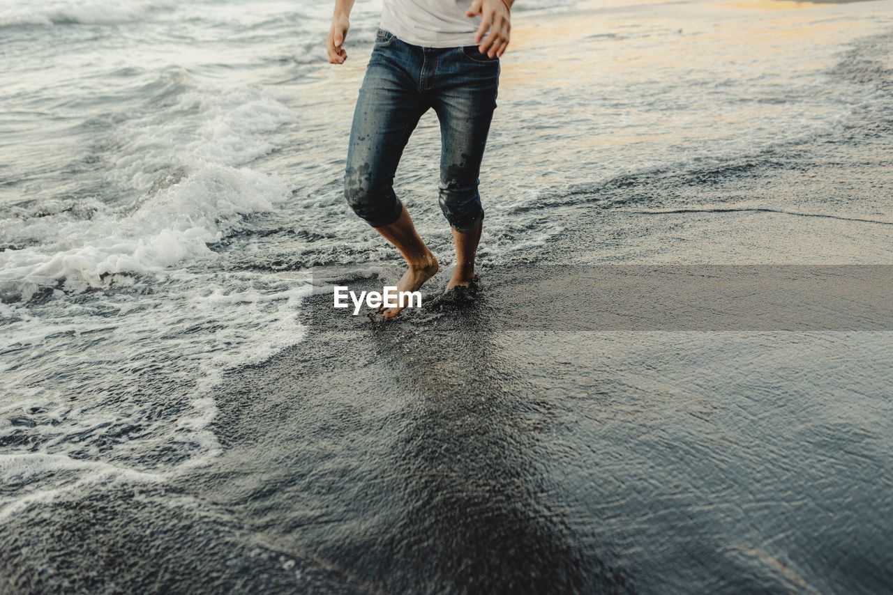 Crop anonymous barefoot male traveler in jeans walking on sandy beach near waving sea at sunset time