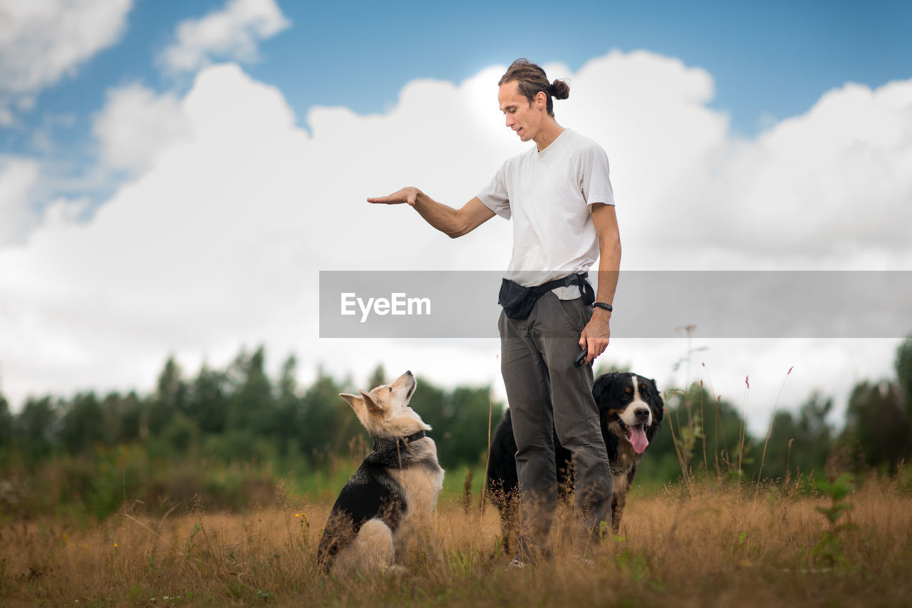 Man playing with dogs while standing on grassy land against cloudy sky