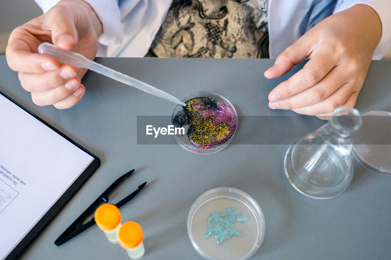 Female scientist adding drop of liquid from from pipette over petri dish with mix of glitters in lab