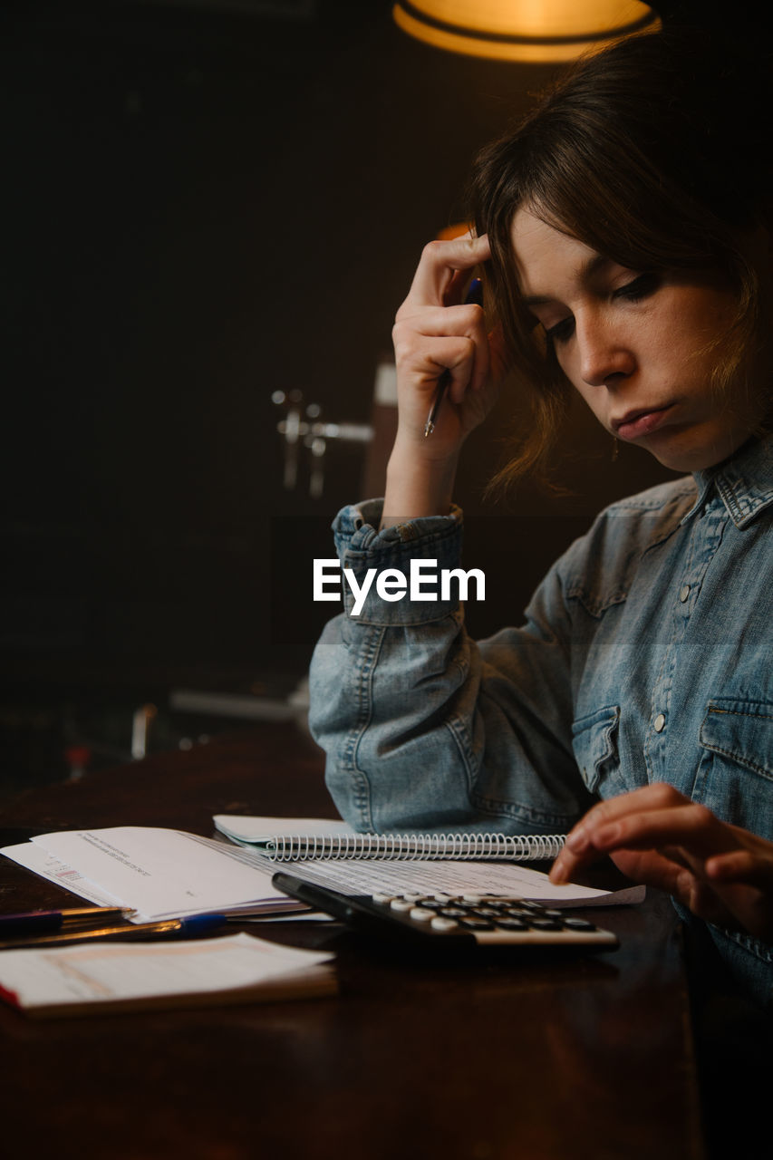 Side view of focused female worker using calculator to count income while sitting at counter with documents during work in cafe