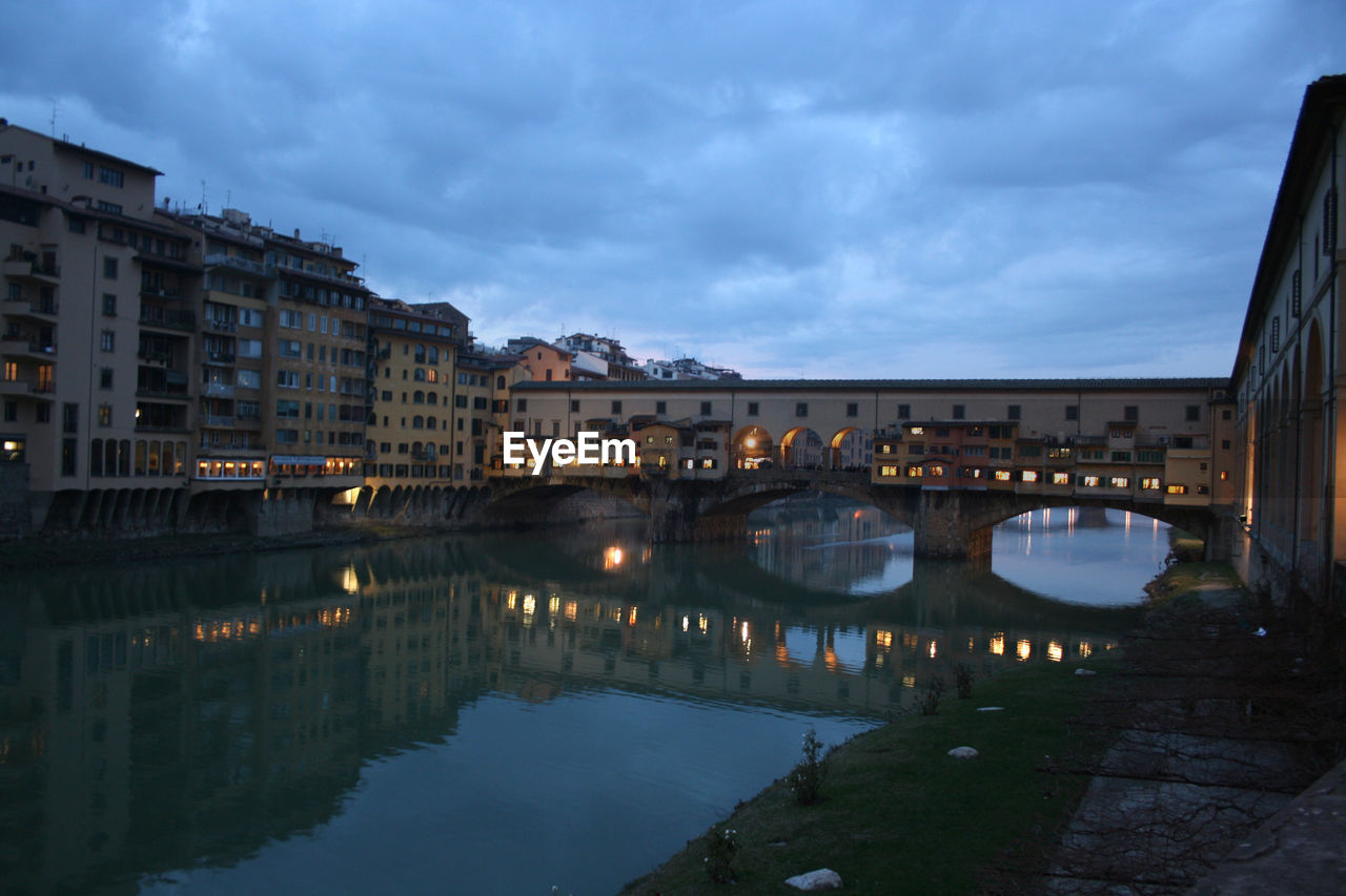 Illuminated bridge over river by buildings against sky at dusk