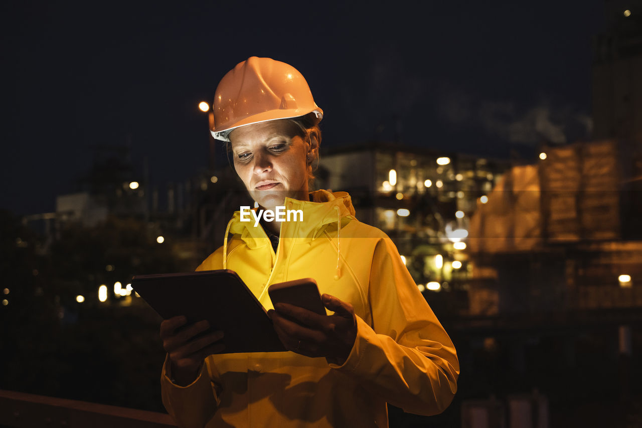 Female professional in yellow hardhat and raincoat using wireless technologies at night