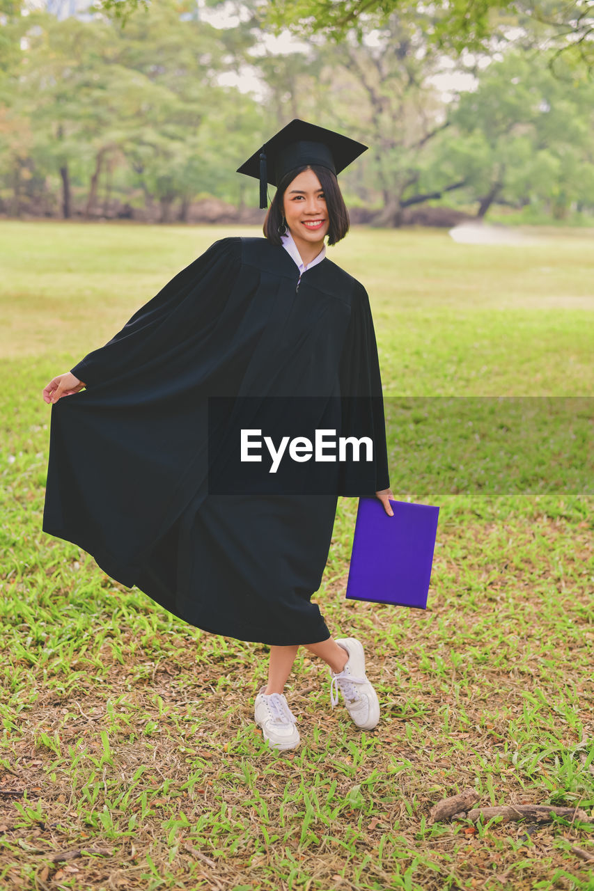 Portrait of young woman in graduation gown holding file while standing at park