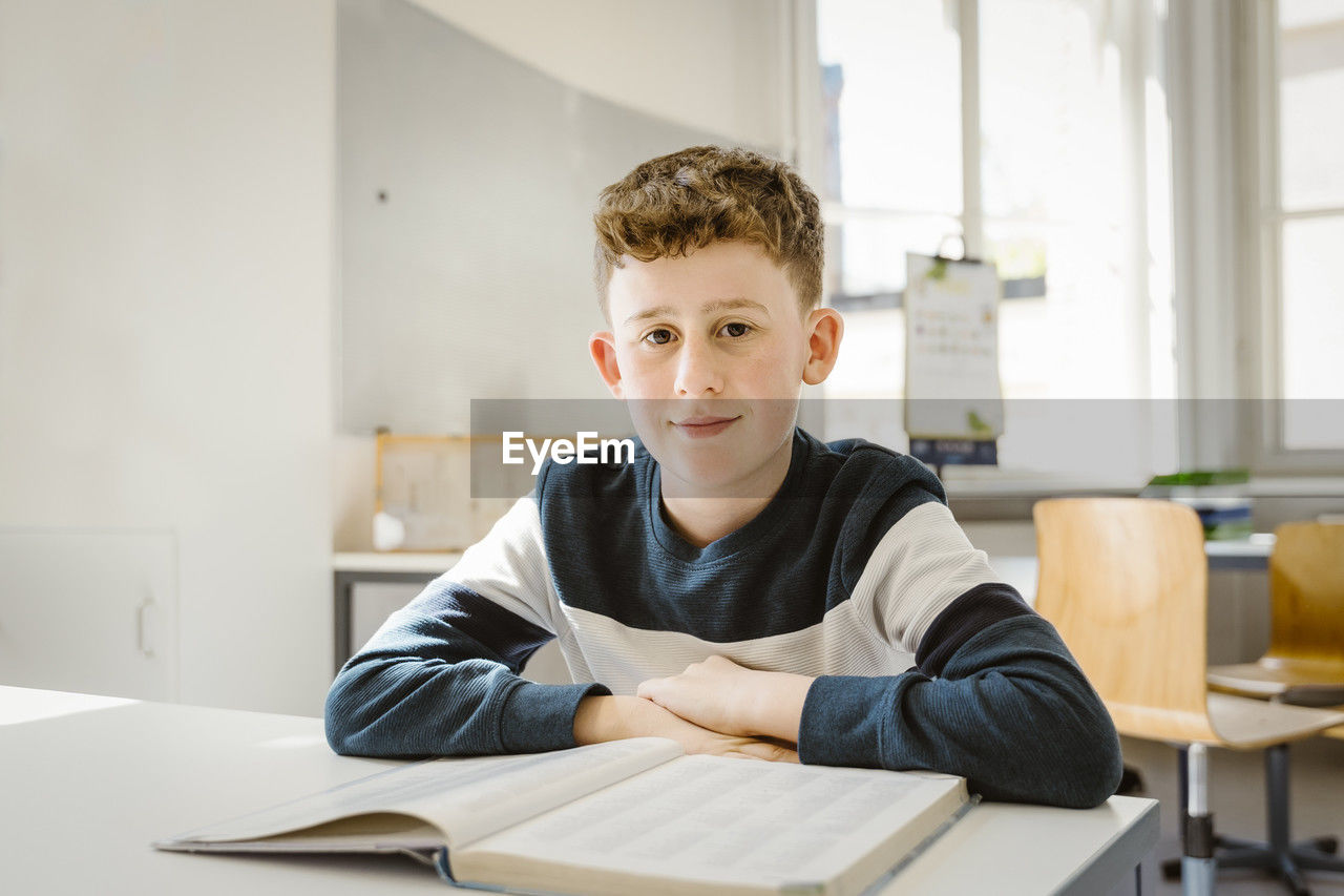 Portrait of schoolboy sitting with book at desk in classroom
