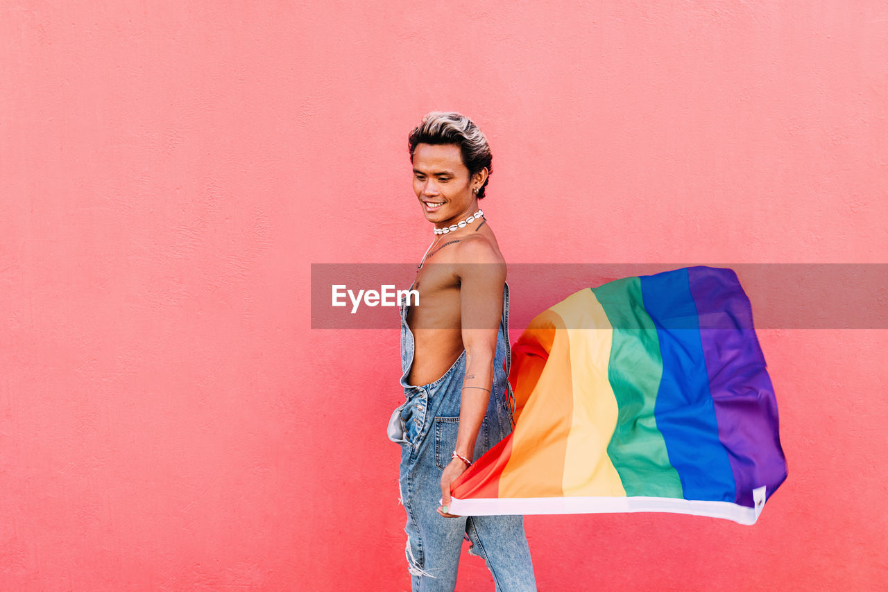 Side view of gay man holding rainbow flag against pink wall