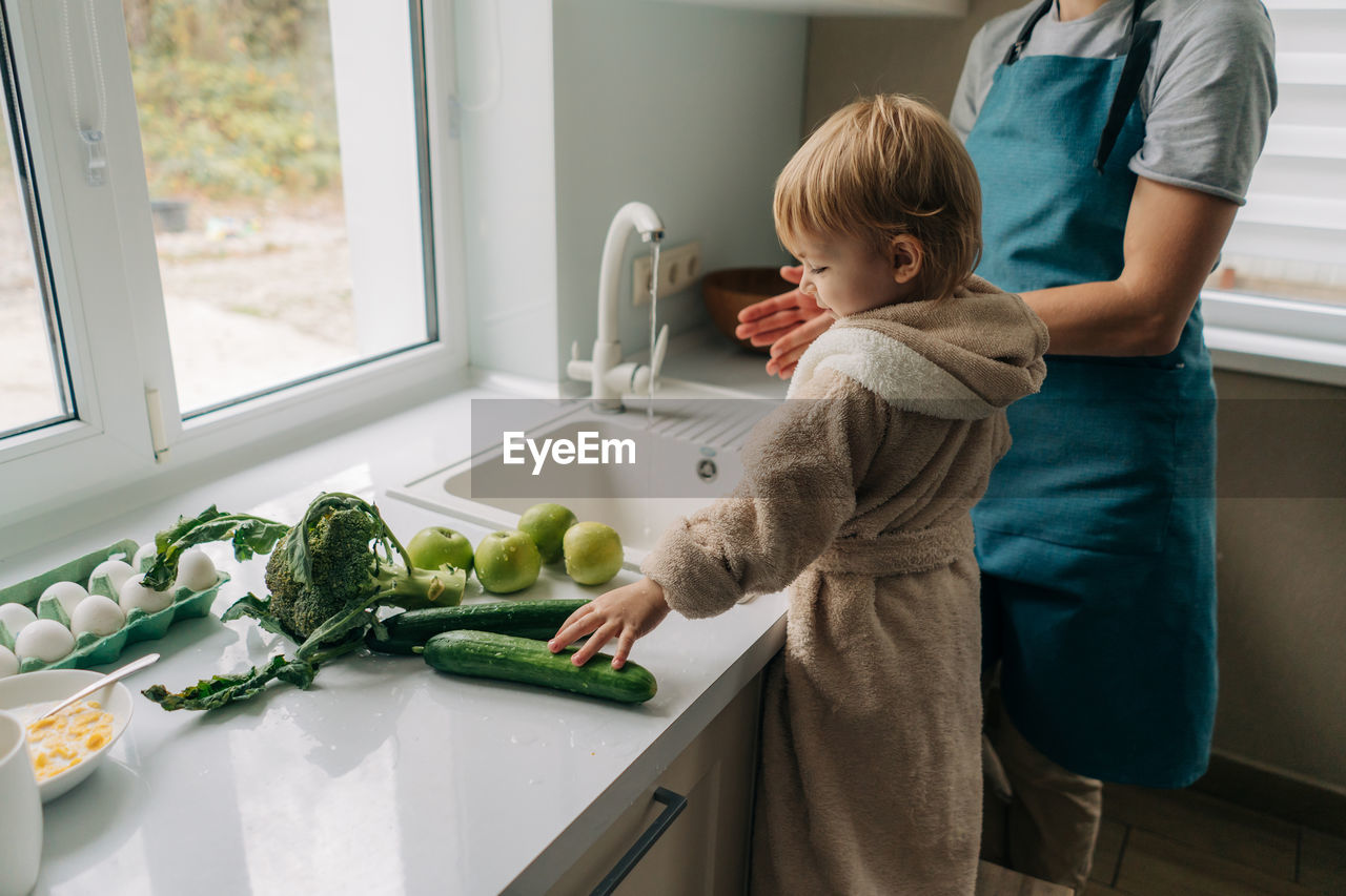 Mother and little child toddler are washing vegetables together in the kitchen for cooking dinner.