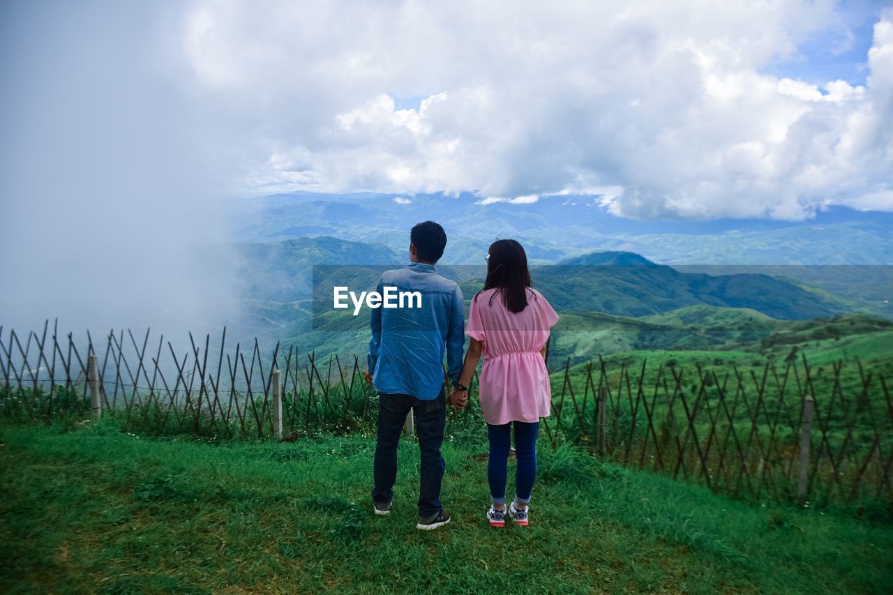 Rear view of man and woman holding hands while standing on grass against sky