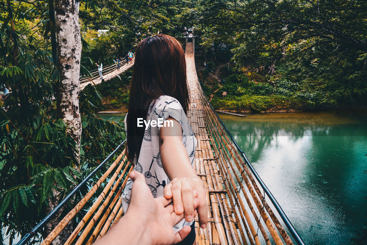 Cropped hand of man holding woman on bamboo footbridge over river in forest