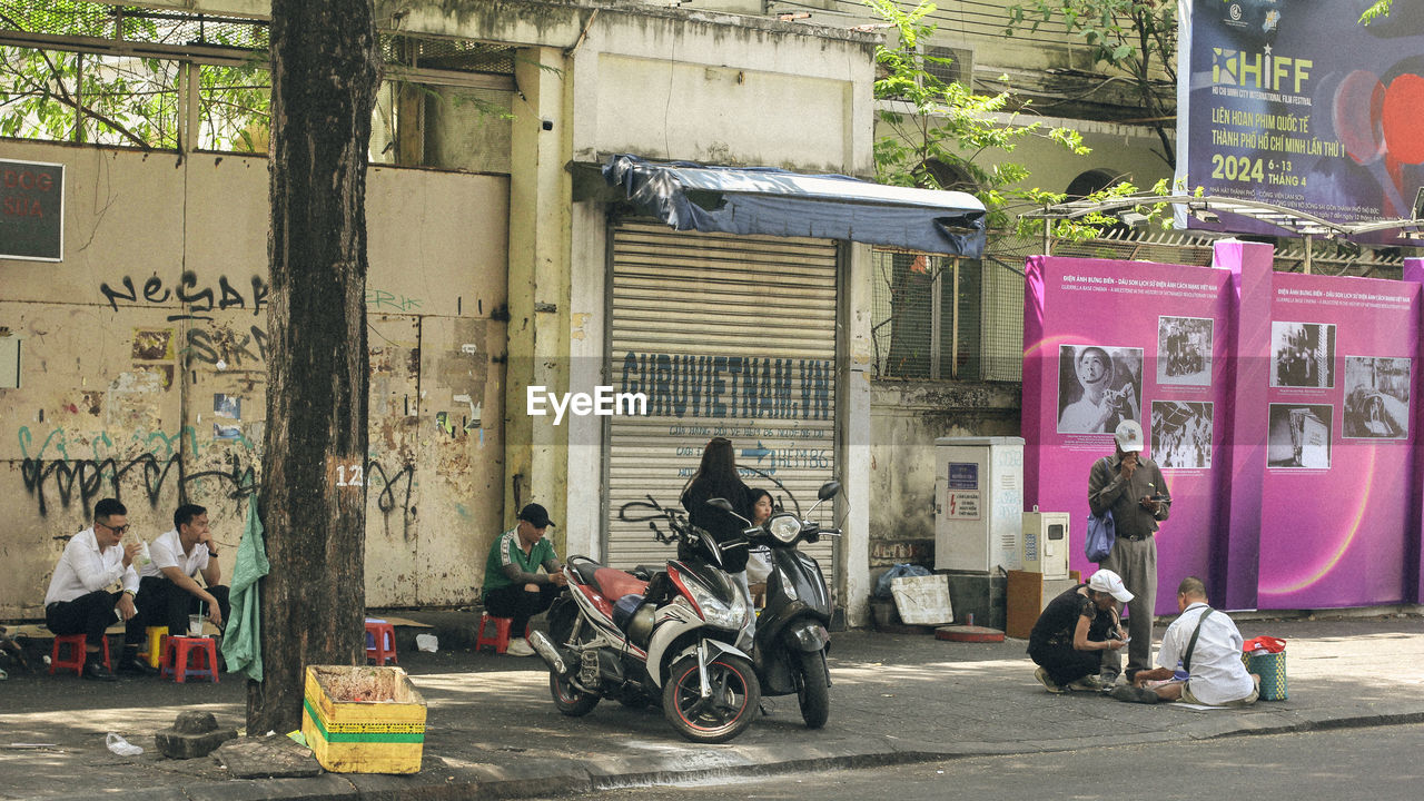 street, road, architecture, city, building exterior, urban area, transportation, built structure, mode of transportation, motorcycle, infrastructure, day, scooter, motor scooter, vehicle, land vehicle, city life, building, outdoors, men, person, group of people, lifestyles, city street, adult, neighbourhood, travel, women, full length, text
