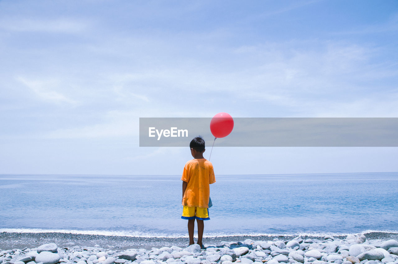 Rear view of boy with red helium balloon standing on shore at beach against sky