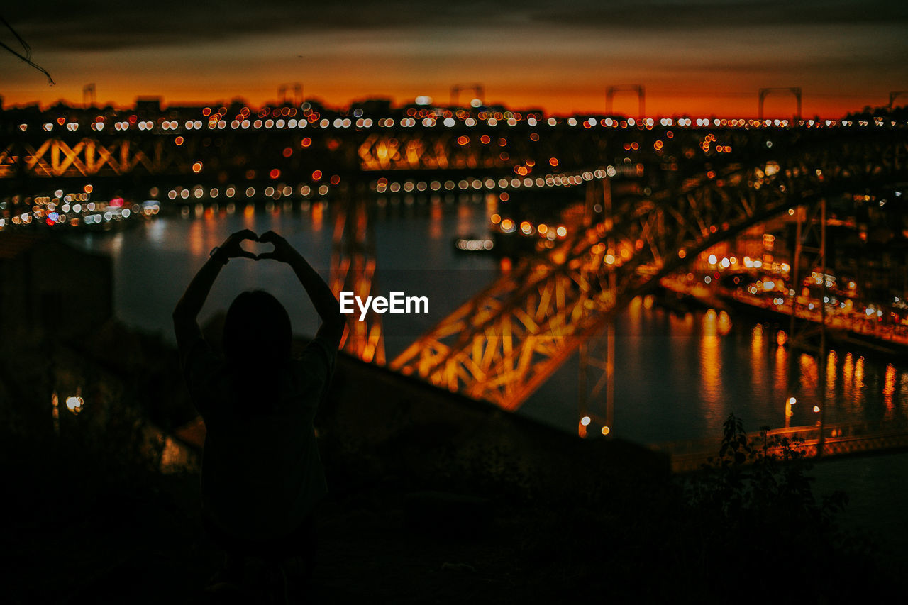 REAR VIEW OF MAN ON ILLUMINATED BRIDGE OVER RIVER AGAINST SKY