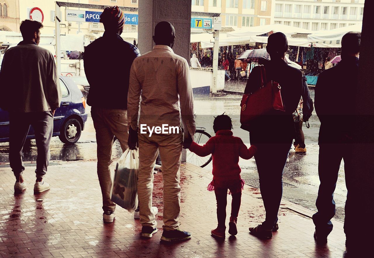 Rear view full length of people standing on sidewalk in city during rainy season