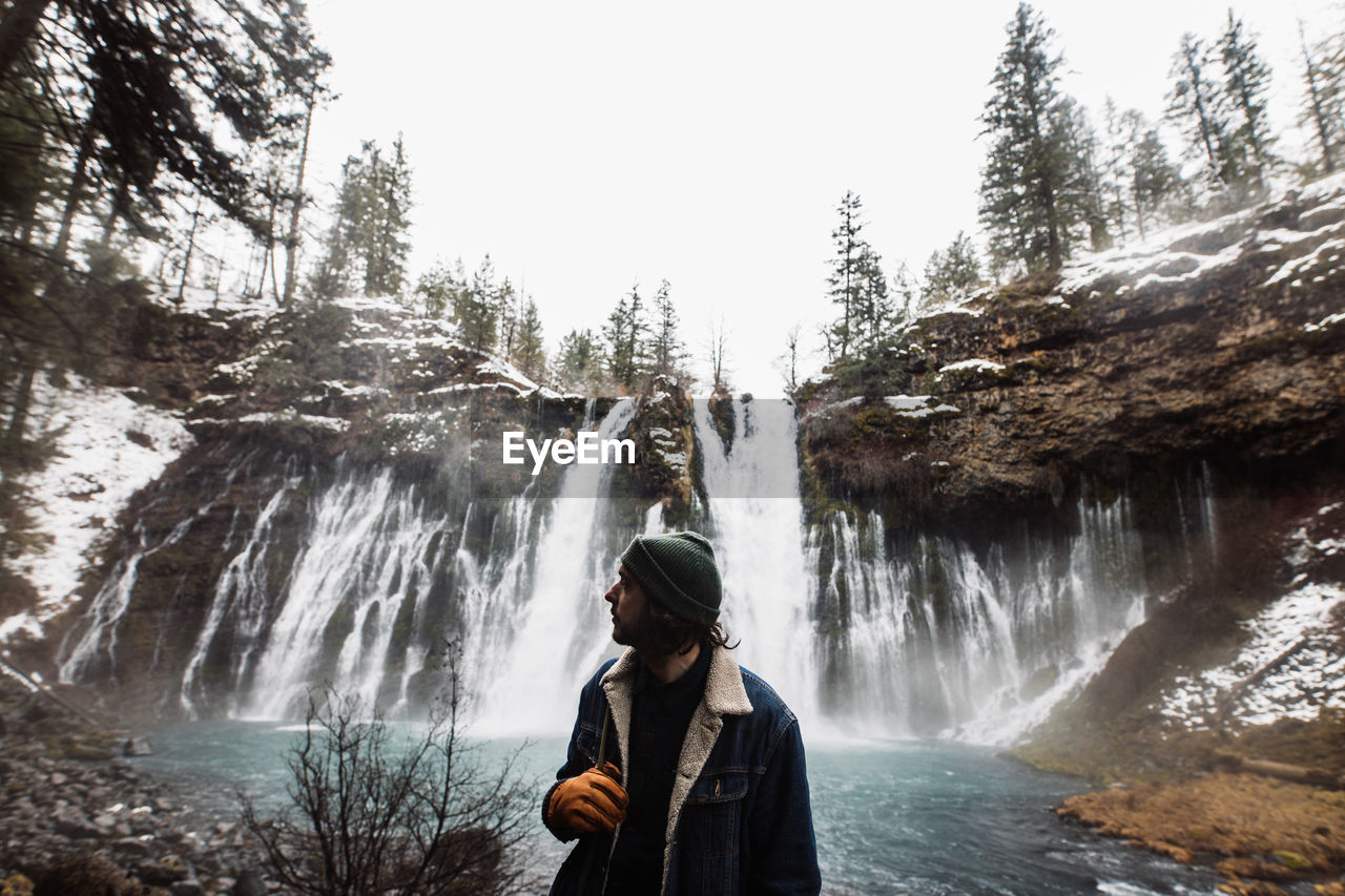 Man standing on picturesque scenery of powerful waterfall with pool flowing among snowy forest in mountainous terrain in winter day in usa looking away