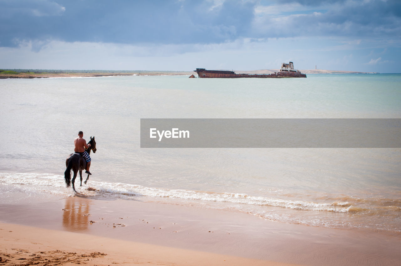 Shirtless man riding horse at beach against cloudy sky during sunny day