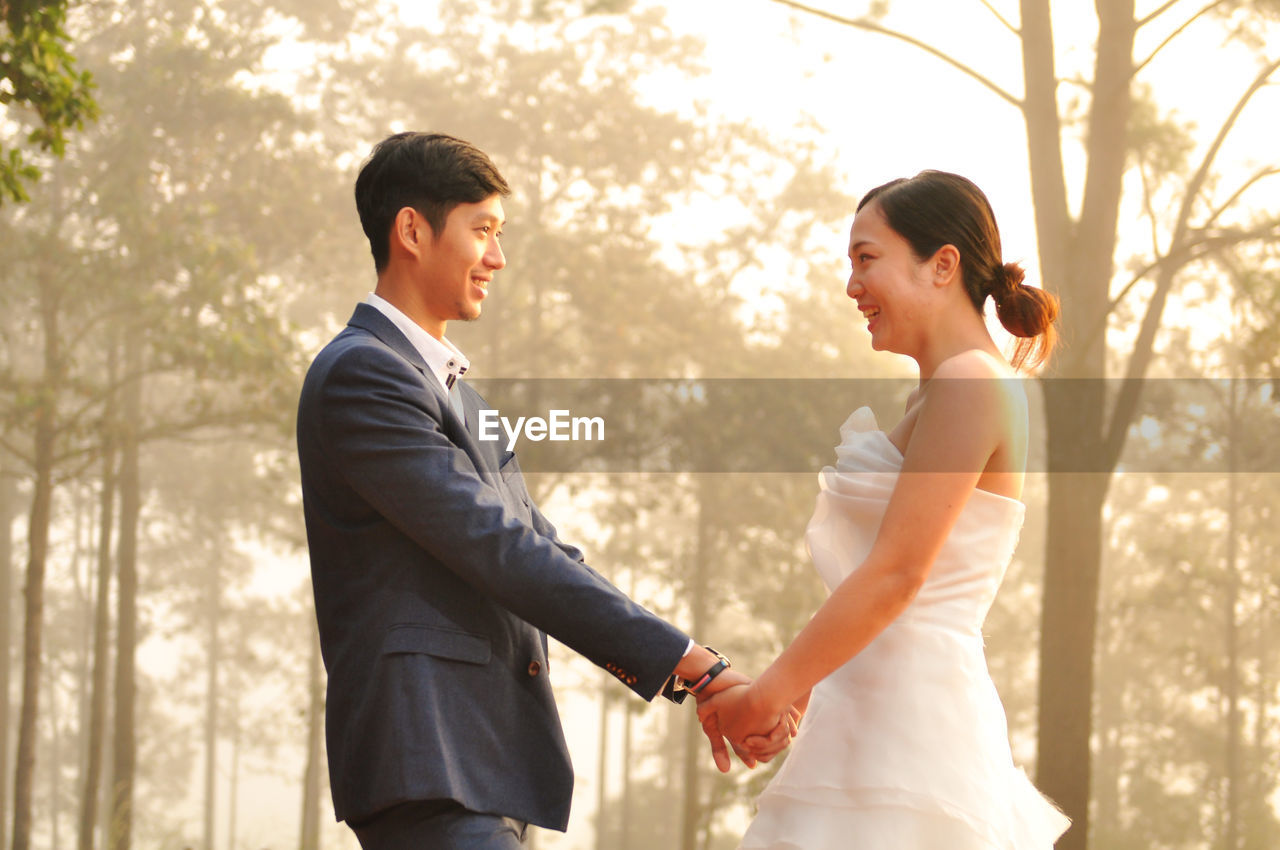 Side view of bride and bridegroom standing against trees