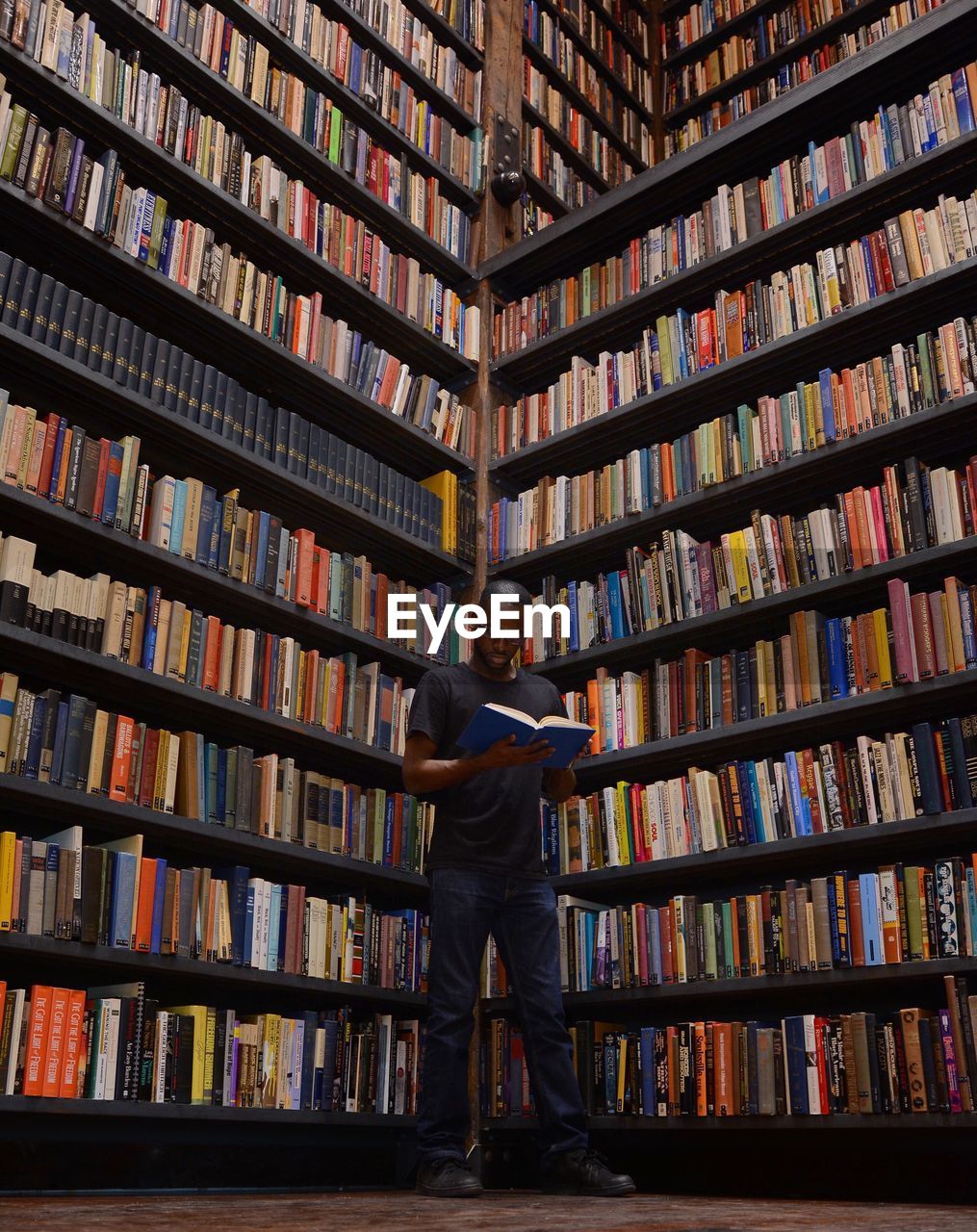 Low angle view of man reading book while standing by bookshelves in library