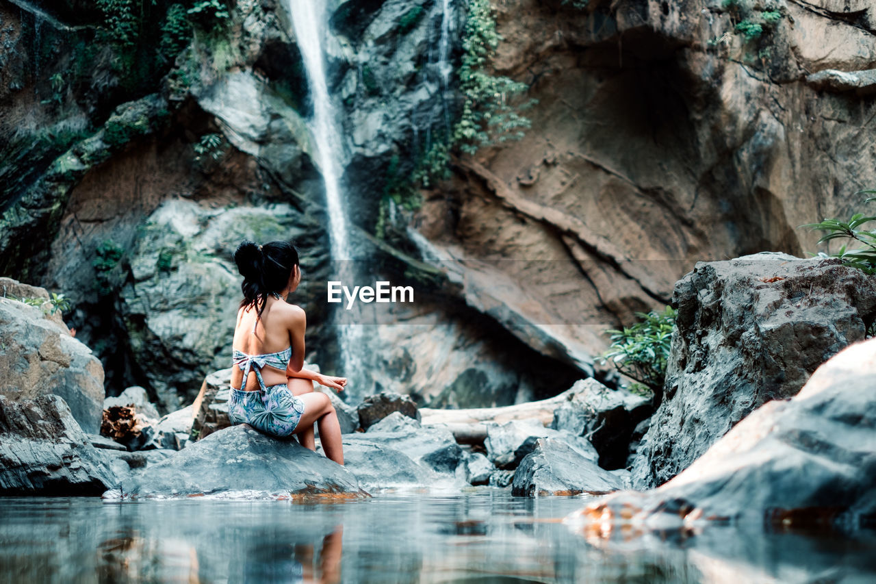 Rear view of woman sitting on rock near waterfall