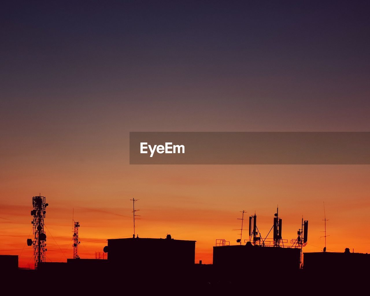 Silhouette antennas over buildings against sky during sunset