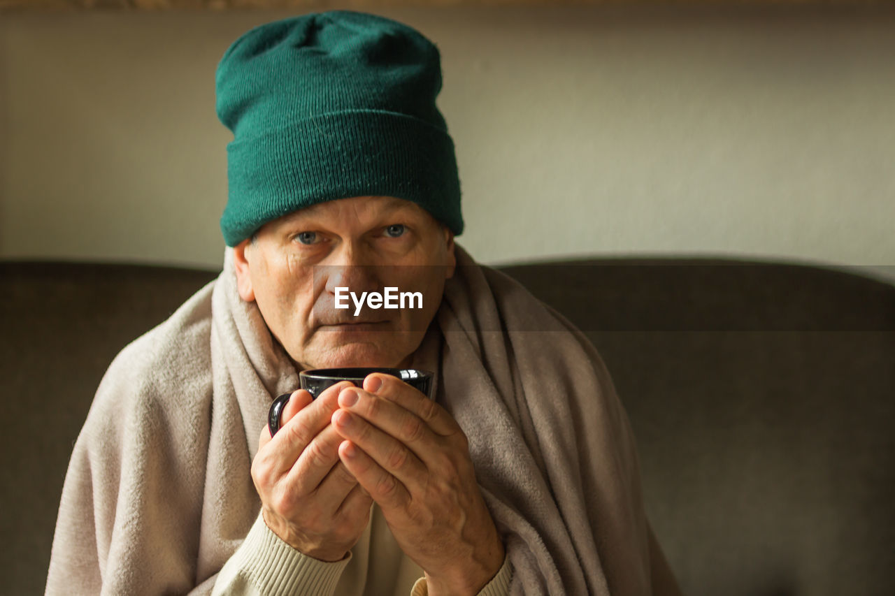 Senior man in green cap wrapped in blanket and drinking hot cup of tea, feeling cold at home.