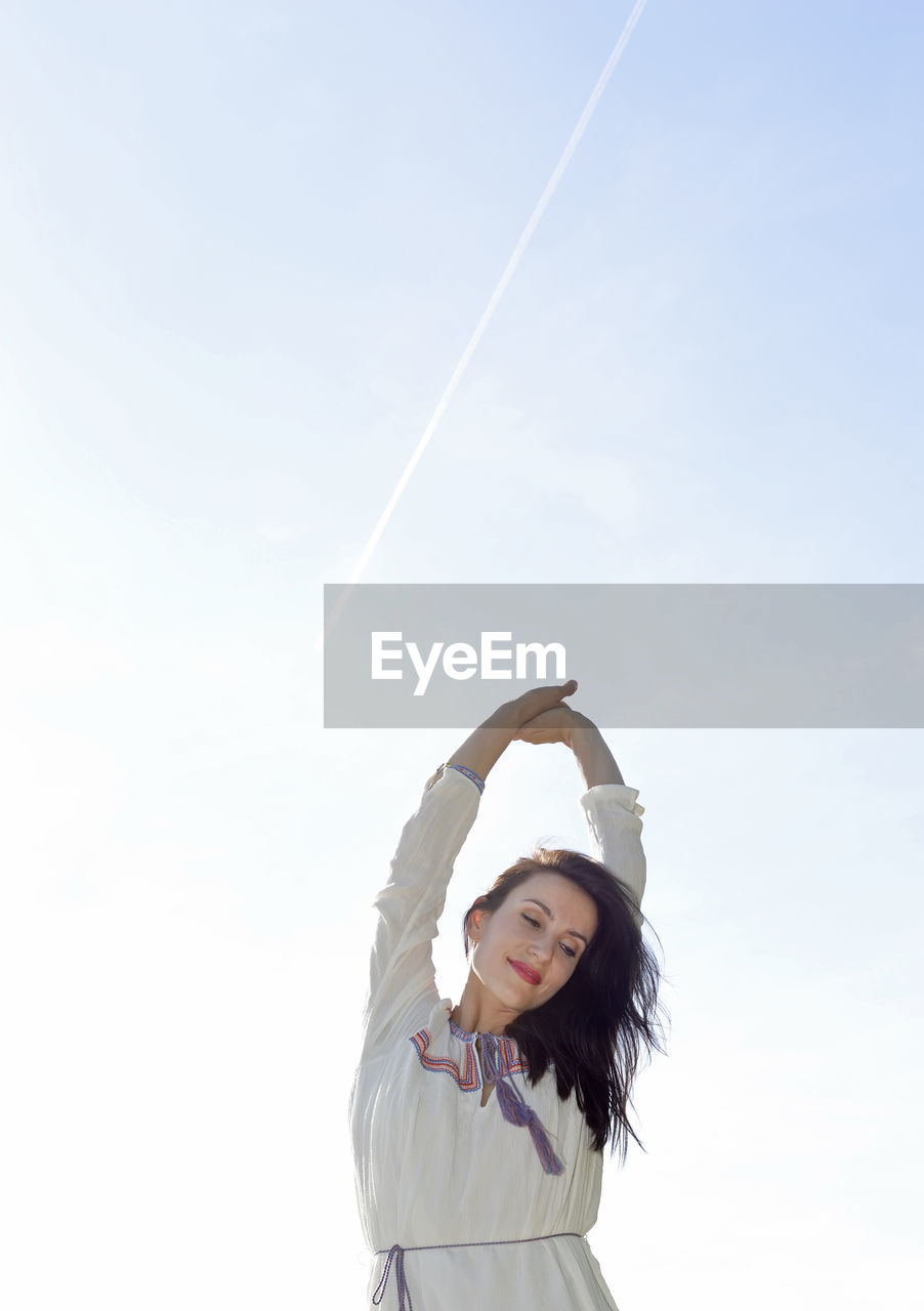 Smiling woman with arms raised standing against clear sky