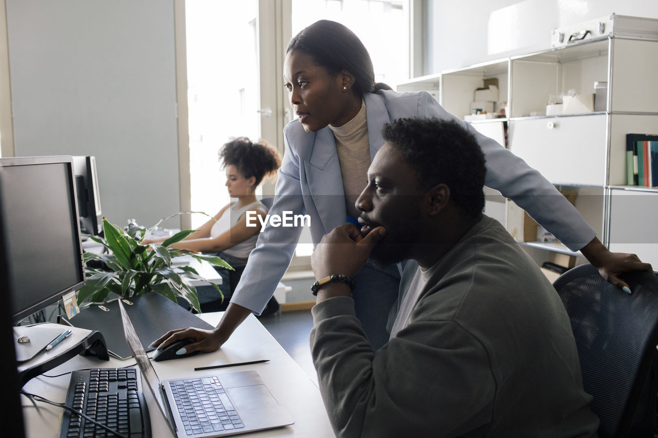 Male colleague with hand on chin sitting near female colleague assisting at office