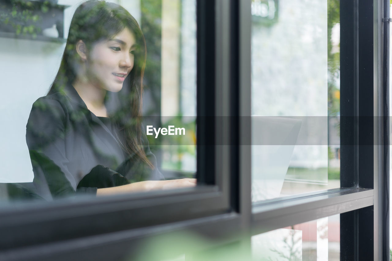 Businesswoman using laptop while sitting in office seen through window