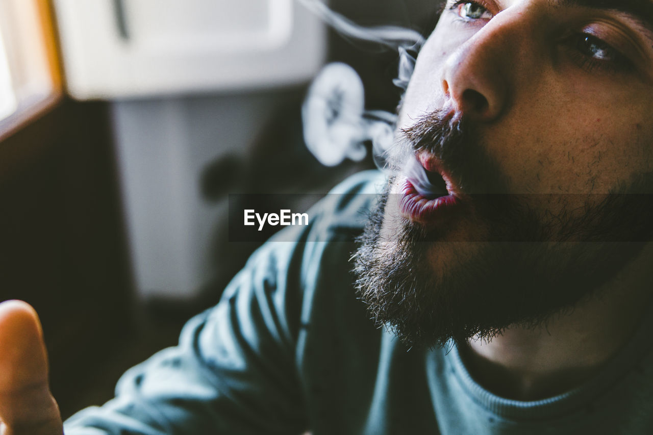 Close-up of bearded man exhaling smoke while smoking marijuana joint at home