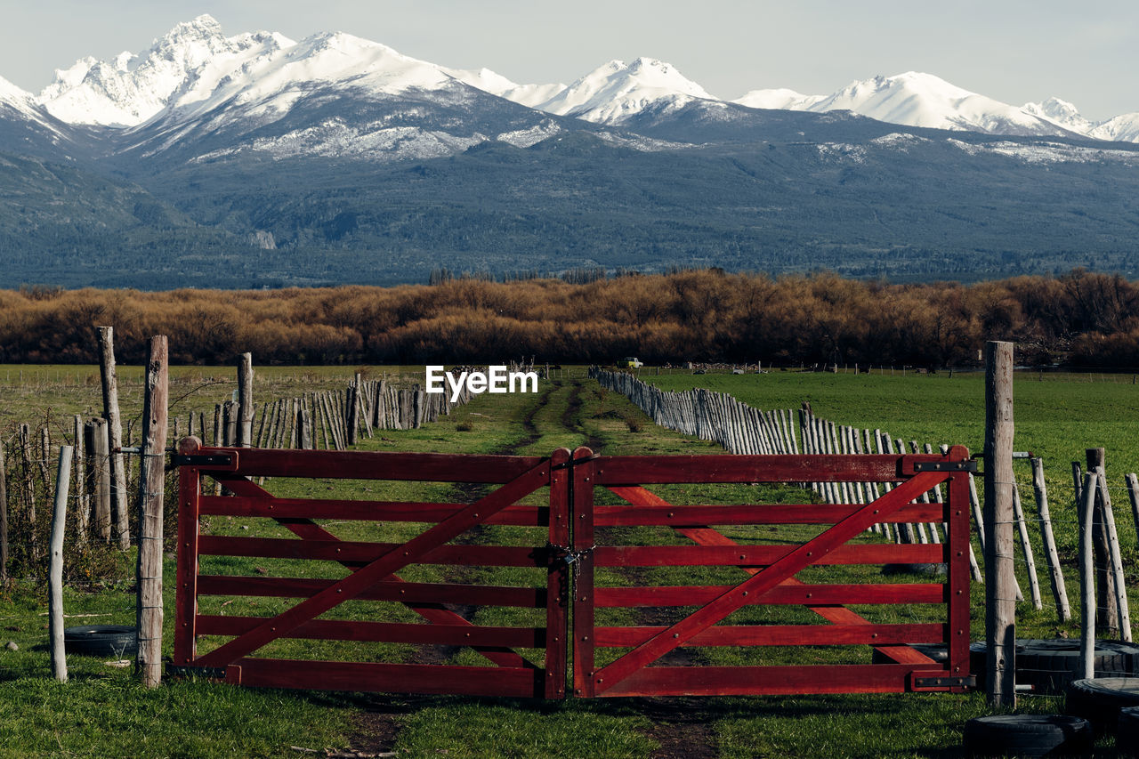 Spectacular scenery of rural road behind shabby wooden fence and gates in valley on background of mountain range covered with snow in patagonia