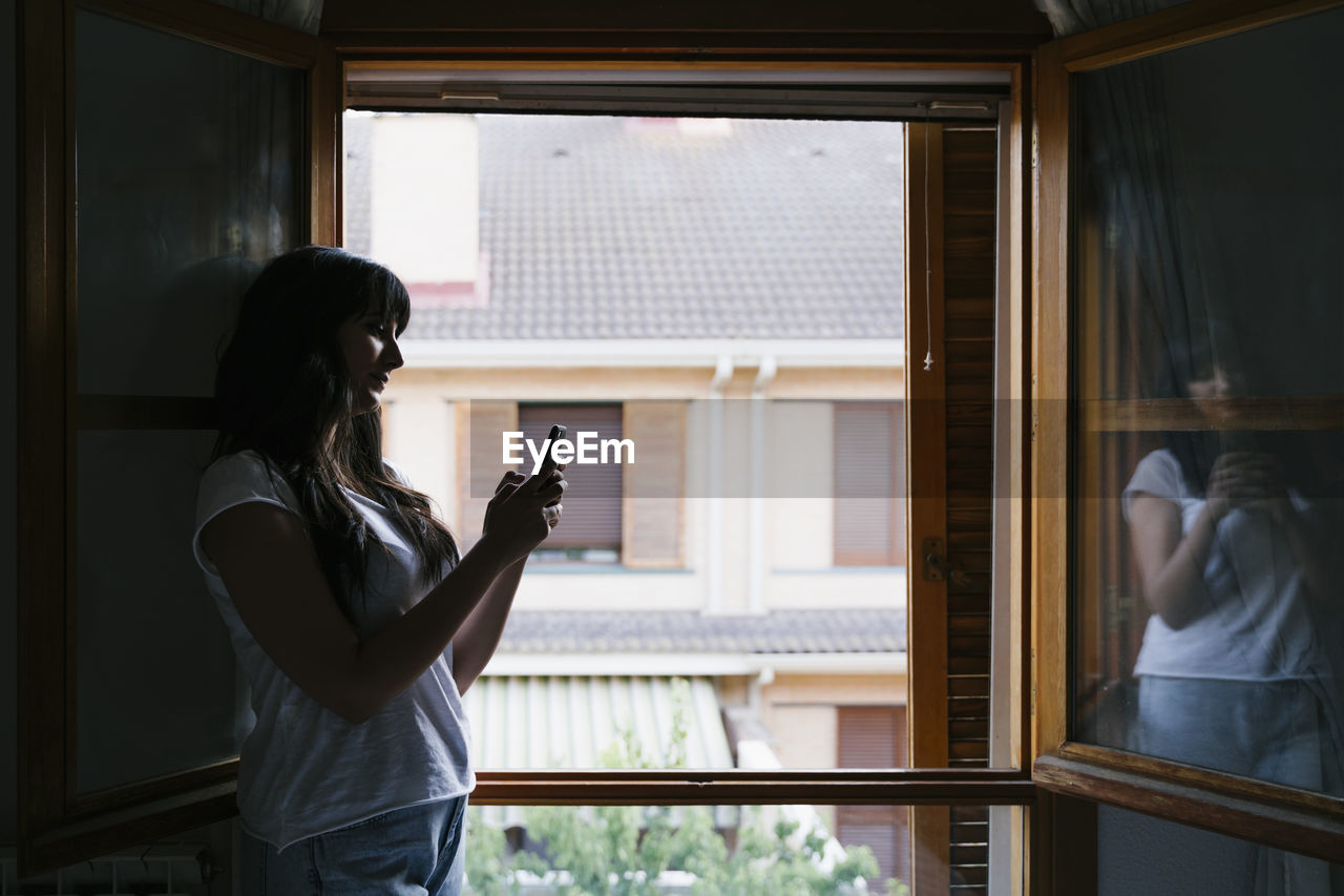 Young woman using mobile phone while standing by window at home