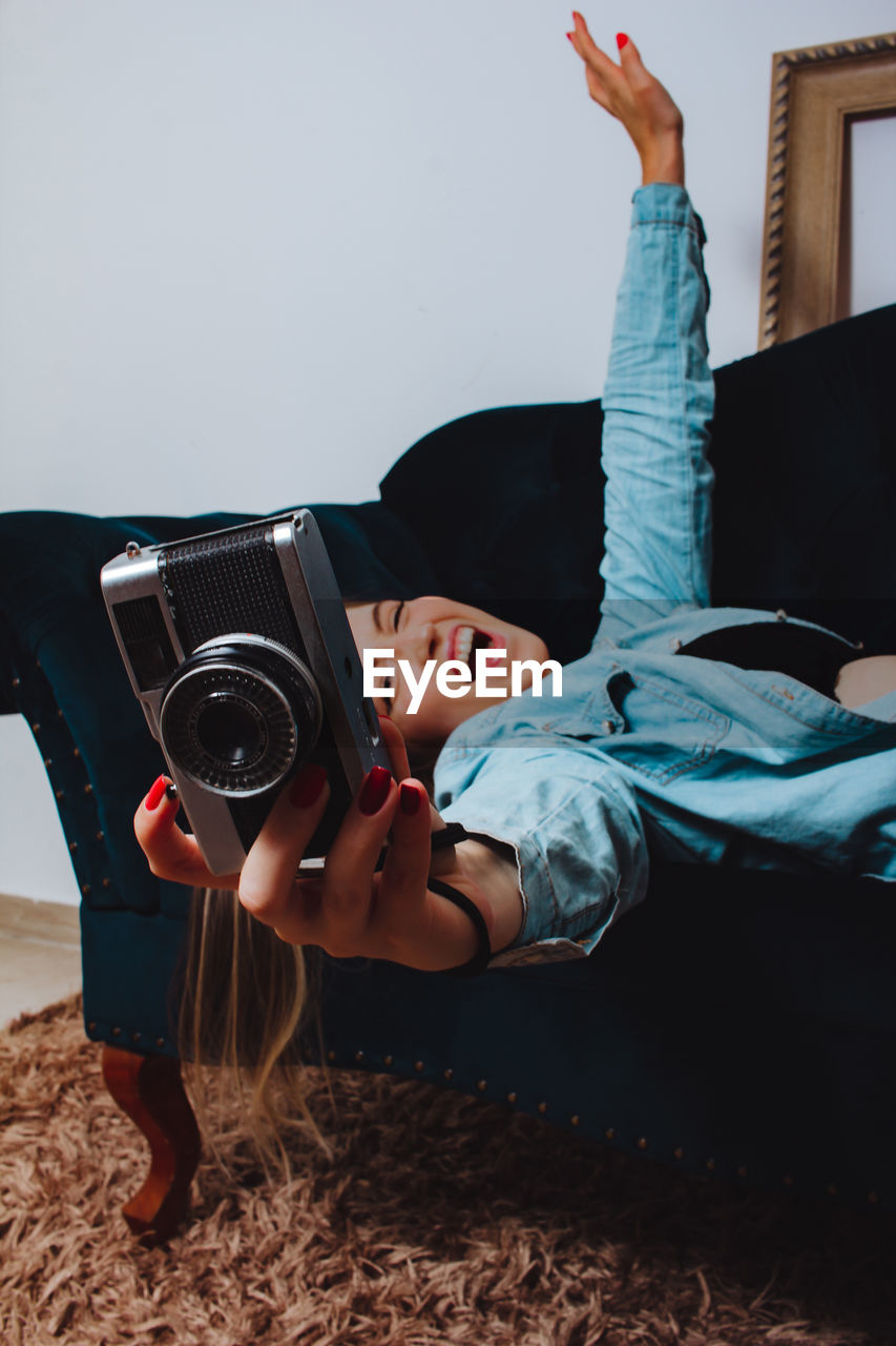 Cheerful woman holding camera lying down on sofa at home